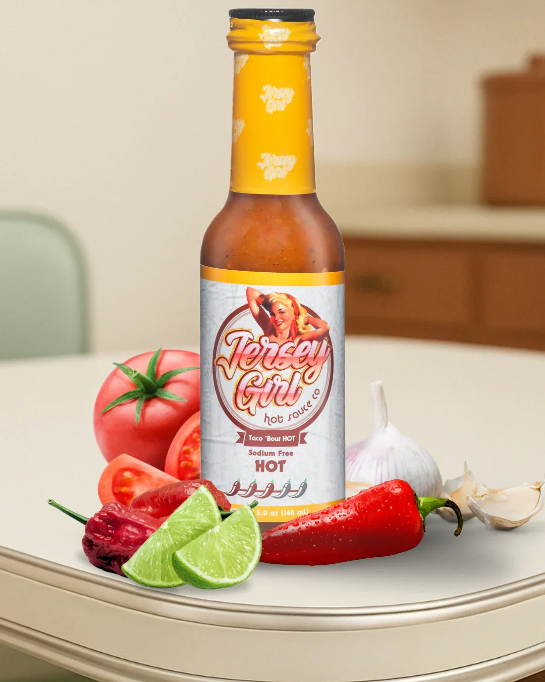 Jersey Girl Taco 'Bout HOT Sauce bottle displayed on a kitchen counter, surrounded by fresh ingredients including lime wedges, garlic, tomato, red chili peppers, and a ghost pepper. The label highlights it as a sodium-free hot sauce with a 3/5-pepper heat rating.