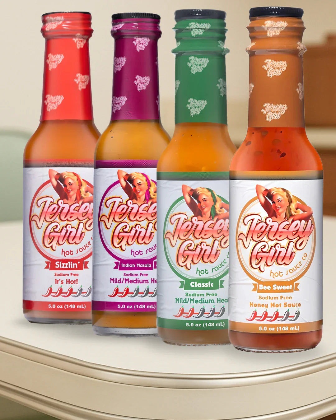 Four bottles of Jersey Girl Hot Sauce displayed in a row on a kitchen counter. From left to right: Sizzlin’ (red label), Indian Masala (purple label), Classic (green label), and Bee Sweet (white label with honey tones). All bottles are labeled as sodium-free, each showcasing a different heat level and flavor profile.