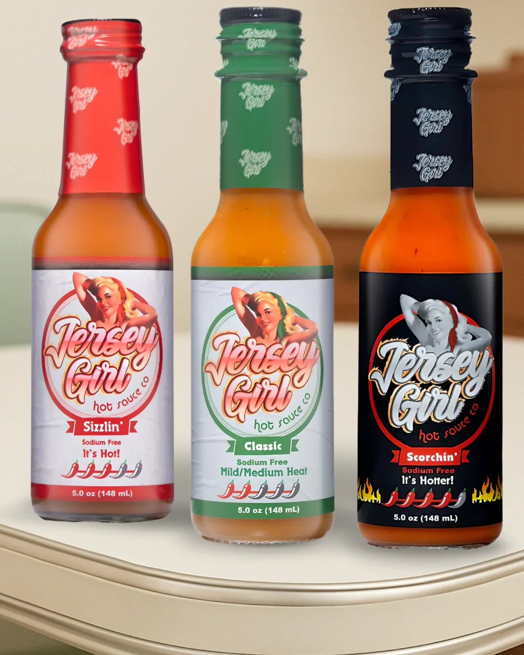 Three bottles of Jersey Girl Hot Sauce displayed on a kitchen counter. From left to right: Sizzlin’ (red label, hot), Classic (green label, mild/medium heat), and Scorchin’ (black label, extra hot with Reaper and Dragon’s Breath peppers). All bottles are 5 oz and sodium-free, showcasing a clean-label design with bold vintage-inspired branding.