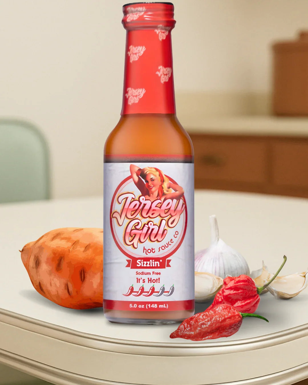 A bottle of Jersey Girl Sizzlin’ Hot Sauce is displayed on a kitchen counter with ingredients beside it, including a sweet potato, garlic cloves, and a red pepper. The label highlights that the sauce is sodium-free and very hot, featuring a vintage-style pin-up design and a 3 pepper heat rating.