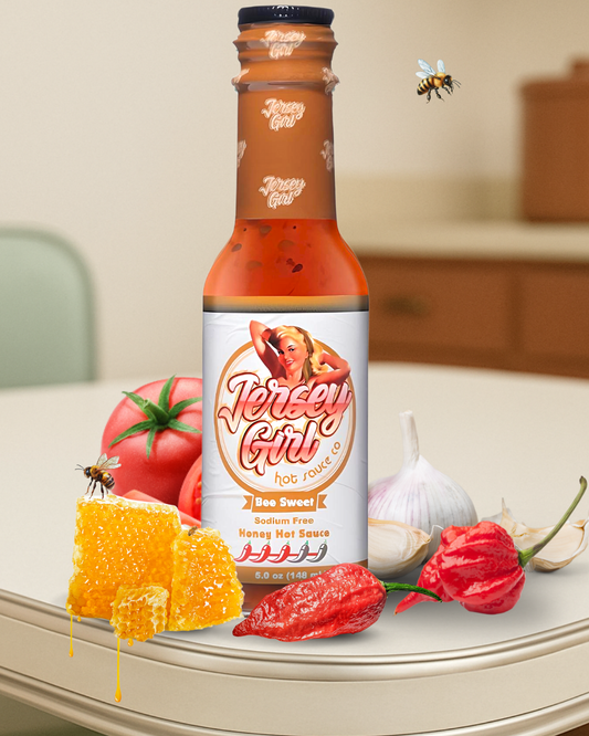 Jersey Girl Bee Sweet Honey Hot Sauce bottle on a kitchen counter, surrounded by honeycomb, tomatoes, garlic, and ghost and scorpion peppers. Bees hover near the bottle, emphasizing the wildflower honey base. The label highlights it as sodium-free with a medium heat level.