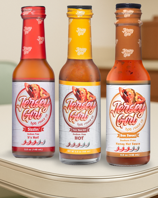 Three bottles of Jersey Girl Hot Sauce displayed on a kitchen counter. From left to right: Sizzlin’ (red label), Taco 'Bout HOT (yellow label), and Bee Sweet (white label with orange accents). Each bottle is 5 oz, sodium-free, and features a heat scale with bold, clean-label branding.