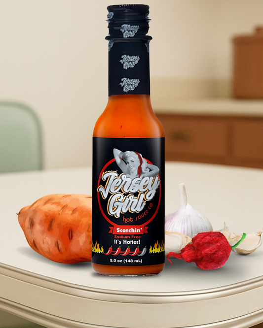 Jersey Girl Scorchin’ Hot Sauce bottle displayed on a kitchen counter, surrounded by fresh garlic, a sweet potato, and a Carolina Reaper pepper. The label highlights it as a sodium-free sauce with intense heat and a 4/5-pepper rating, marketed as “It’s Hotter!”